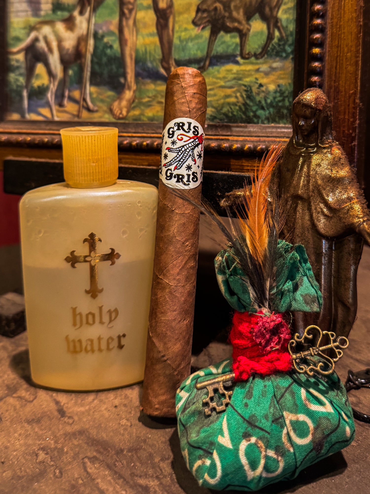 Vintage bottle labeled 'holy water', cigar reading 'Gris Gris',  decorative bag with feathers and keys, and painting of animals in the background.