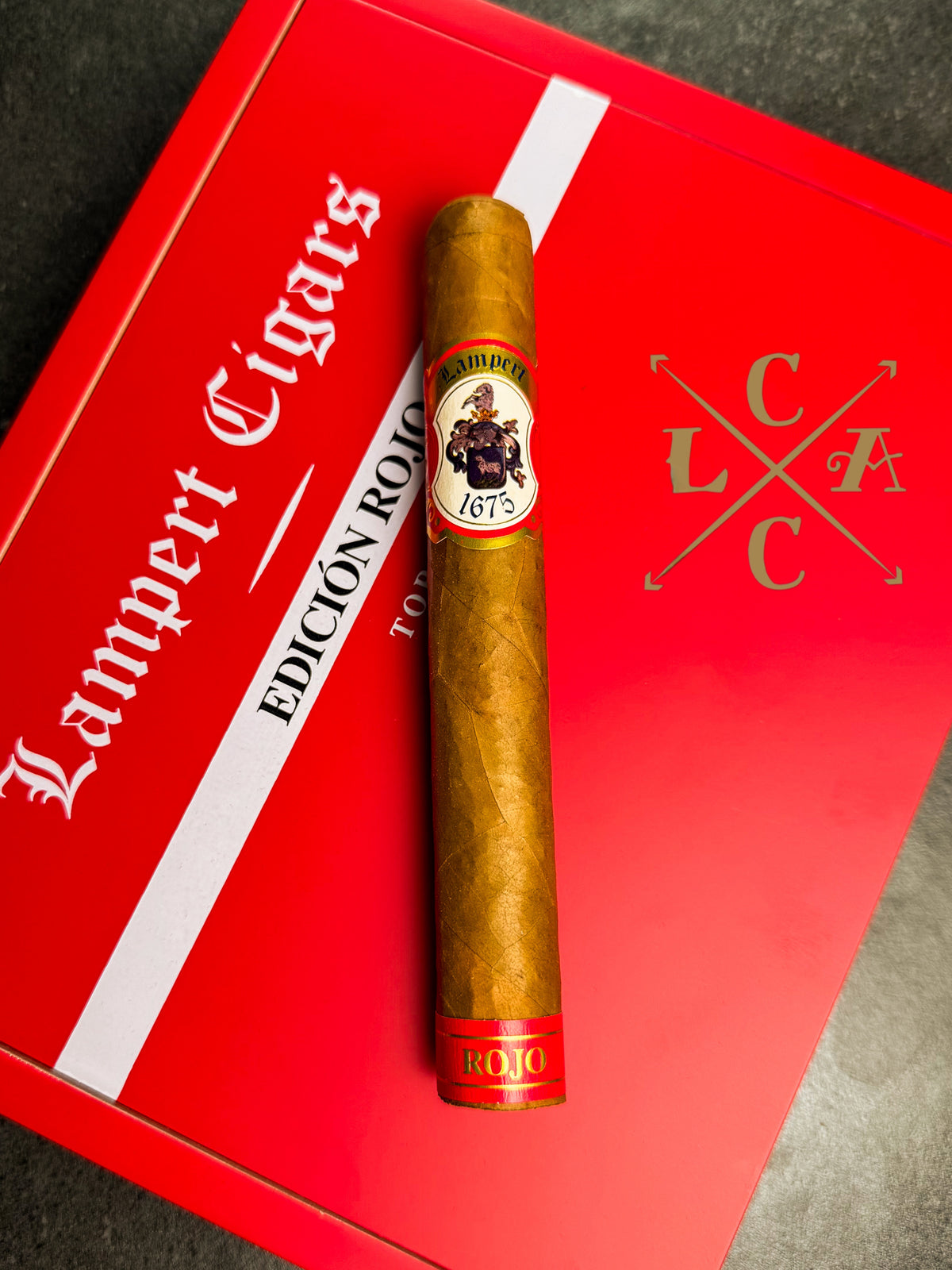 A single gold-brown cigar with a circular '1673' band, displayed vertically on top of a red and white Lampert Cigars box.