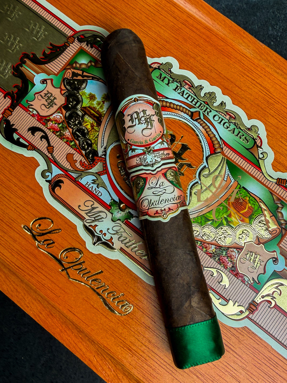 A dark brown cigar with a green tip and a decorative paper band, placed diagonally on an ornate wooden box lid featuring colorful printed artwork and gold script.