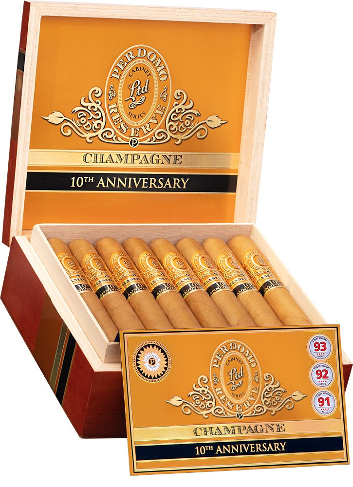 A box of Perdomo Champagne cigars, 10th Anniversary edition, with cigars visible inside the open lid. The exterior is gold with black and gold text, and a wooden box frame is visible.