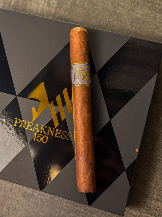 A cigar resting on a black surface with a geometric pattern and the text 'PREAKNESS 150'.
