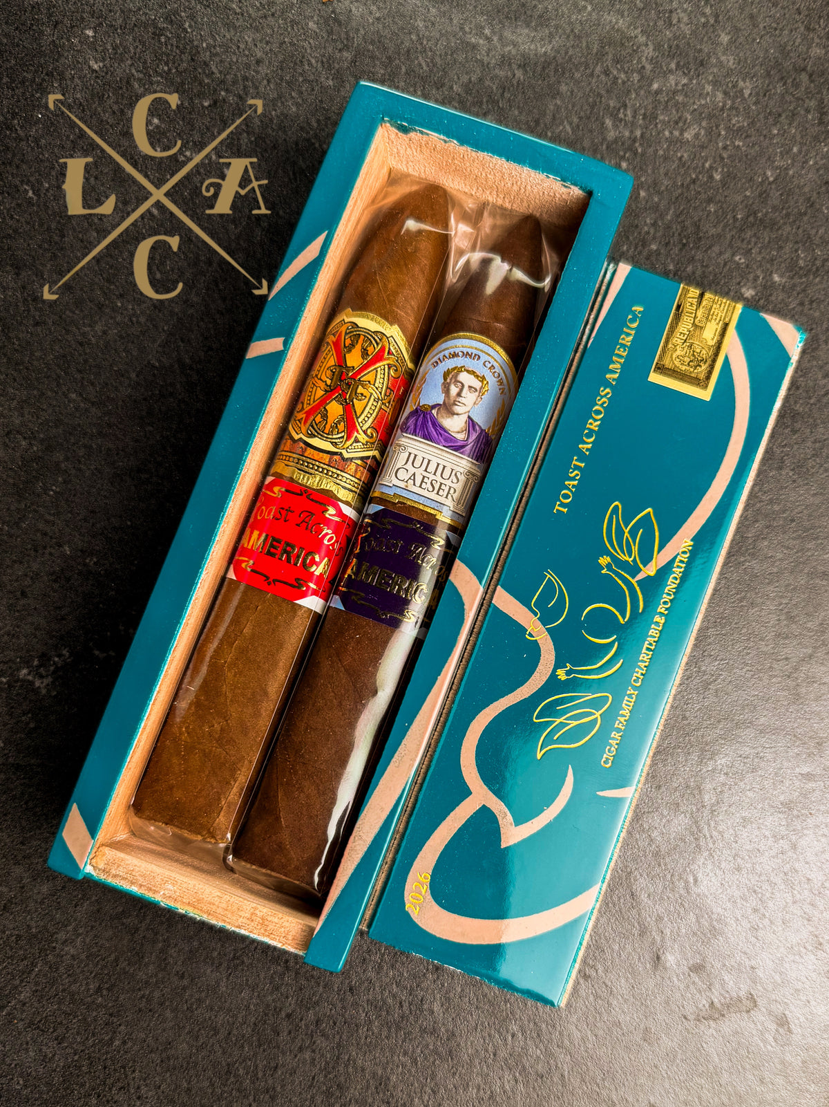 A luxury cigar gift set featuring two cigars in a teal wooden presentation box next to its matching teal lid, displayed on a dark textured slate background.
