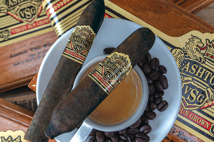 Two cigars on a white plate with coffee beans and a small bowl of honey, surrounded by Ashtray & VSG branded items.