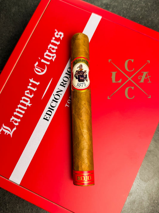 A single gold-brown cigar with a circular '1673' band, displayed vertically on top of a red and white Lampert Cigars box.