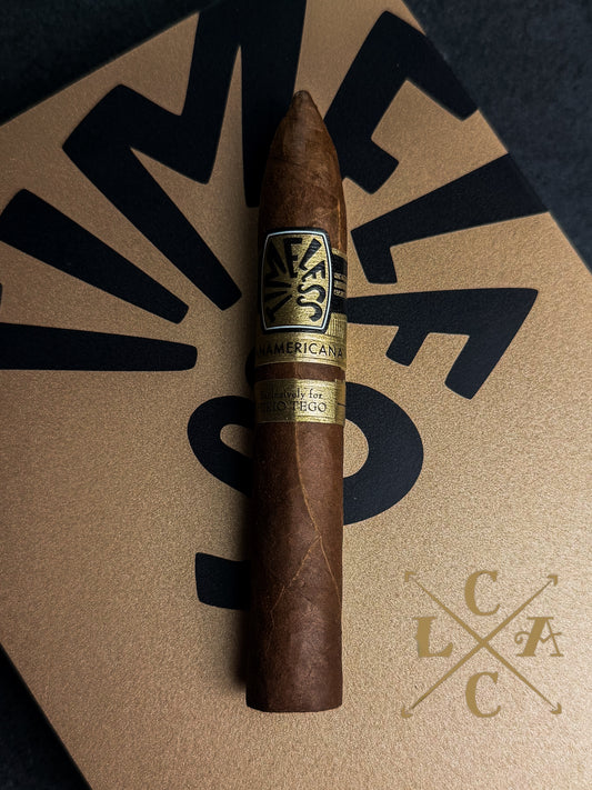 A single cigar placed upright on a tan textured surface with bold black graphic lettering in the background. The cigar has a brown wrapper and a gold-and-black band reading 'P.