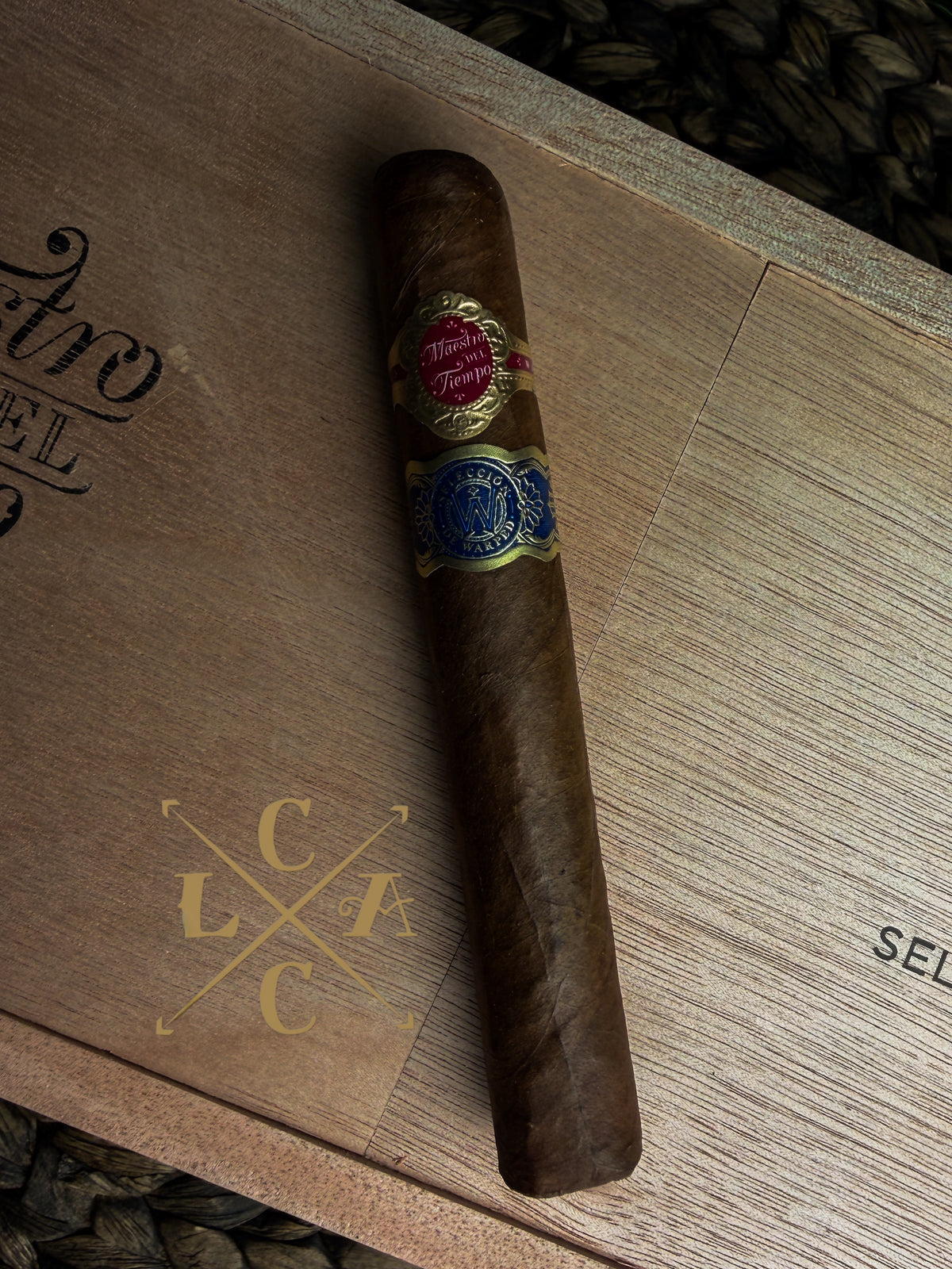 A single dark brown cigar resting diagonally on a wooden box lid, featuring a red and gold wrapper band and a blue and gold band.