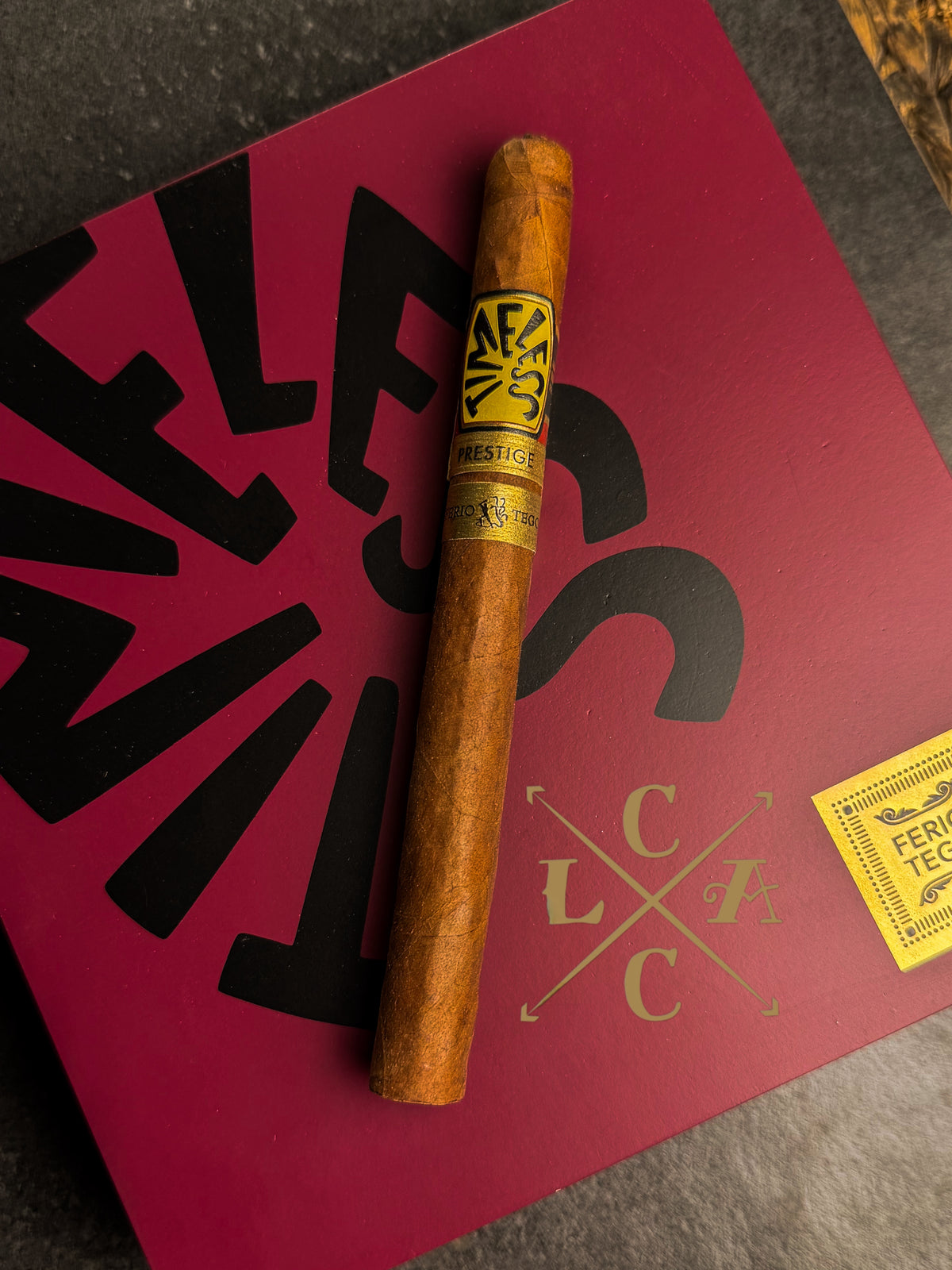 A single cigar resting diagonally on a dark textured surface, placed on top of a red branded box with black lettering and a small yellow sticker. The cigar band shows the brand name AF&S.