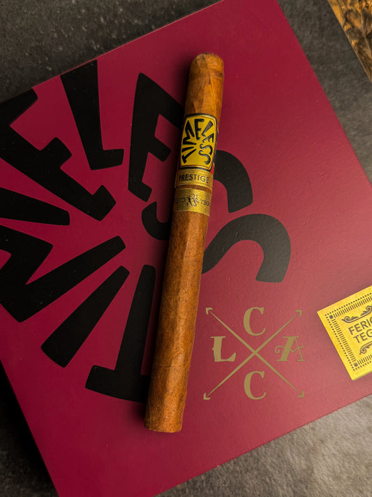 A single cigar resting diagonally on a dark textured surface, placed on top of a red branded box with black lettering and a small yellow sticker. The cigar band shows the brand name AF&S.
